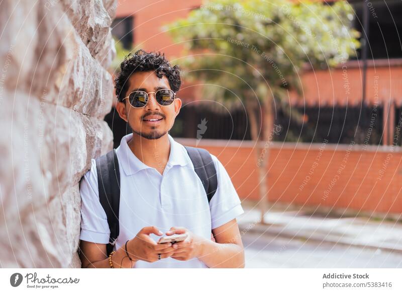 Positive man standing on street stone wall summer style satisfied urban smile sunlight vacation male city hispanic guy sunglasses glad personality lean on shirt