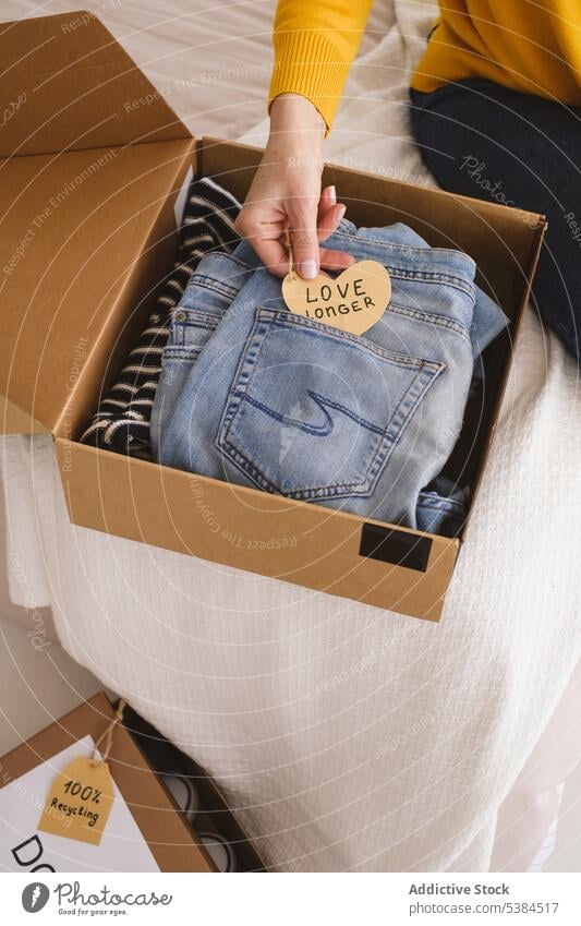 Crop woman packing clothes in cardboard box donation charity donate note gift package jeans carton paper heart pocket casual belonging prepare love denim shape