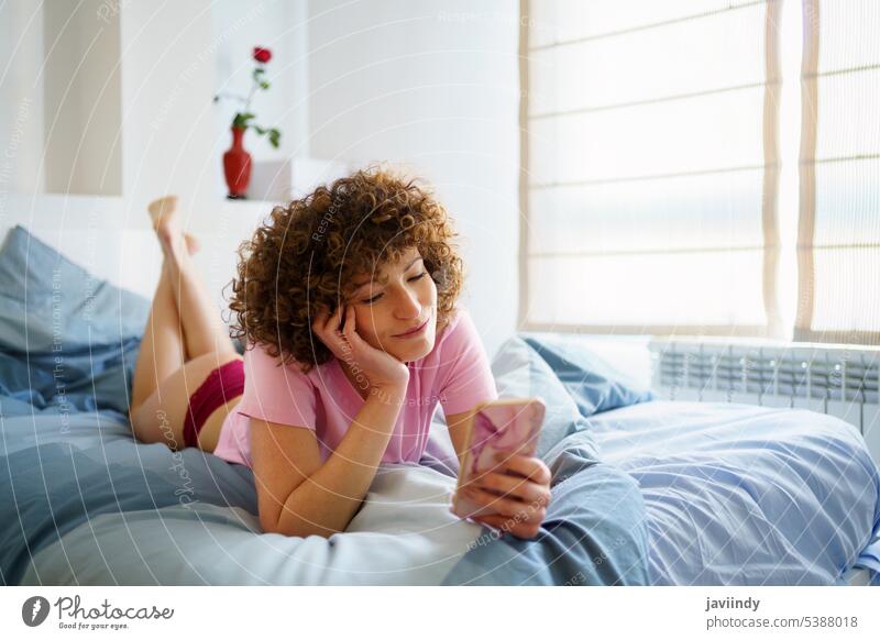 Young woman in love, with curly hair, looking at something beautiful on her smartphone using social media smile message rest comfort bed social network home
