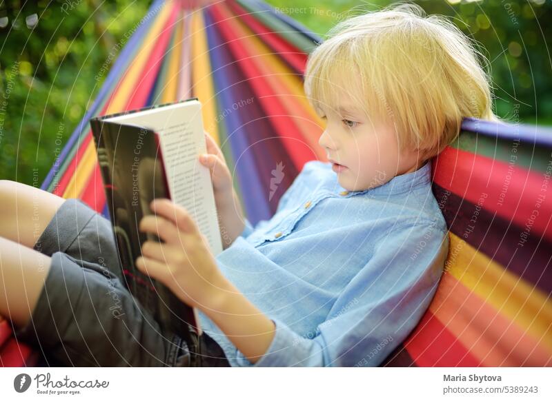 Cute little blond caucasian boy reading book and having fun with multicolored hammock in backyard or outdoor playground. Summer leisure for kids. Child swinging and relaxing in hammock.