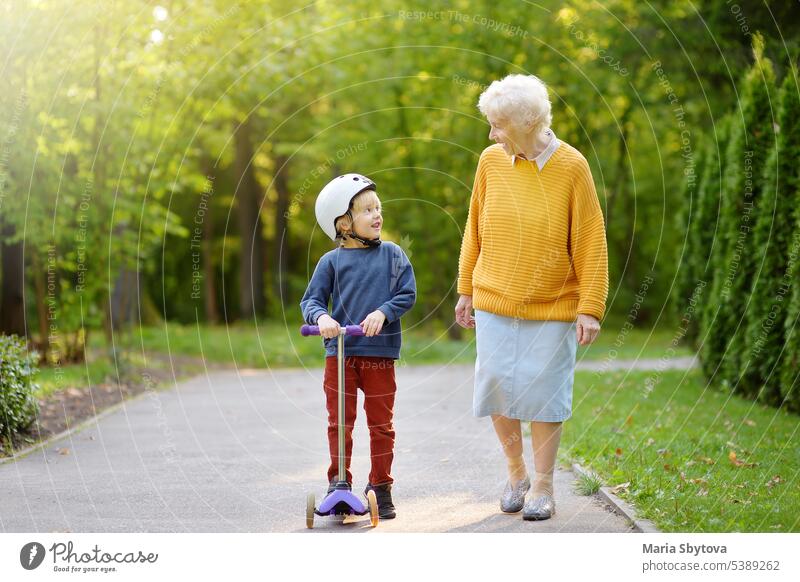 Beautiful granny and her little grandchild walking together in autumn park. Boy riding by scooter. grandma kid grandson elder grandmother family stroll toddler