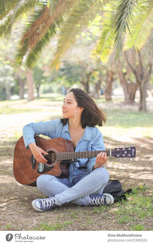 Young woman playing guitar in the park in summer, sitting under the green palm tree. Millennial female musician. leisure beautiful musical performance