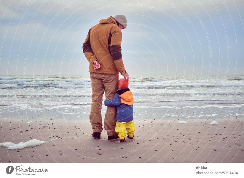 A toddler holds on to a man while he cuddles him reassuringly on the head, beach, waves in the background Father with child Trust Child Affection