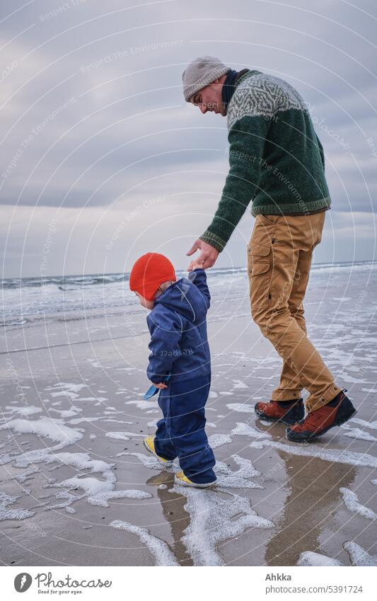 A man in knitted sweater and light pants helps a small child with red cap to explore on the beach first steps Toddler Father Trust Paternity Conduct Support
