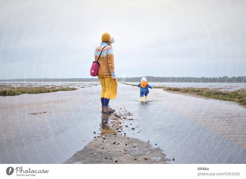 Toddler in colorful rain clothes runs away on flooded street while mother looks behind Flood Gale Mother with child Self-confident Freedom Independence
