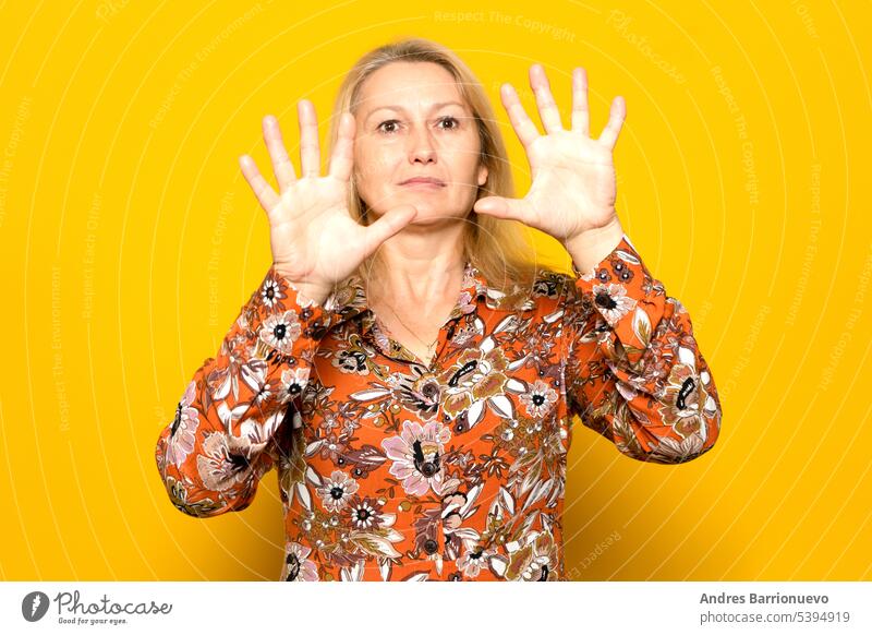 Caucasian blonde woman in her 40s wearing a patterned sweater posing worried with her palms in front of her face, isolated on yellow studio background. people