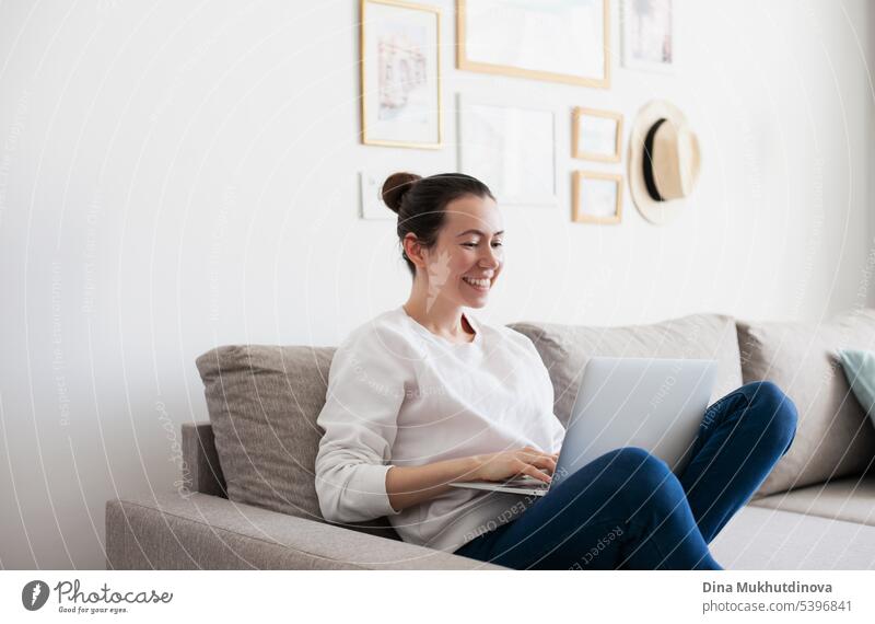 Young woman working on laptop. Millennial girl working remote job from home, using mobile phone, sitting on the cozy sofa, smiling. background beautiful