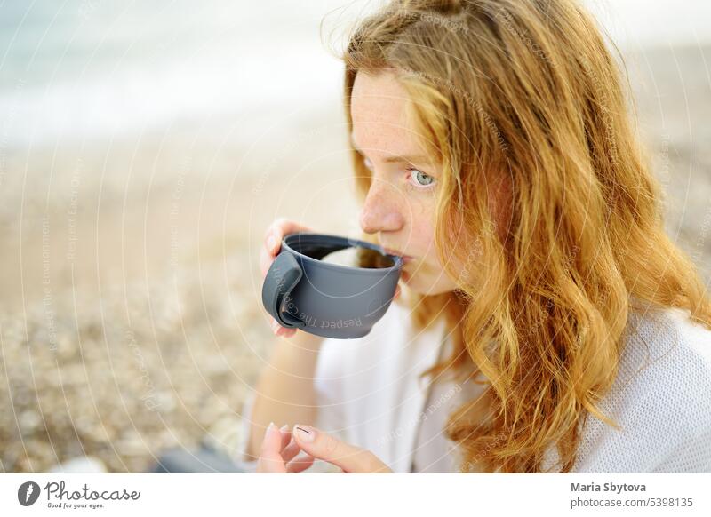 A young redhead woman is having a picnic by the sea shore. hot drink thermos tea beverage adult beach family person hang out people nature white meet happy girl