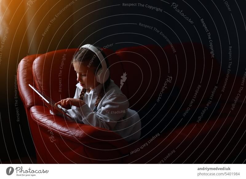 Concentrated teen girl in headphones plays game, communicate on internet social media, listens to music or online studies at home on digital tablet sitting on sofa. Children tech addiction concept