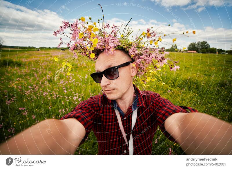 Young smiling man in a wildflower wreath and sunglasses takes a selfie on a wide-angle camera is having fun in the field on summer day. Positive guy doesn't have allergy. Lifestyle summer Concept.