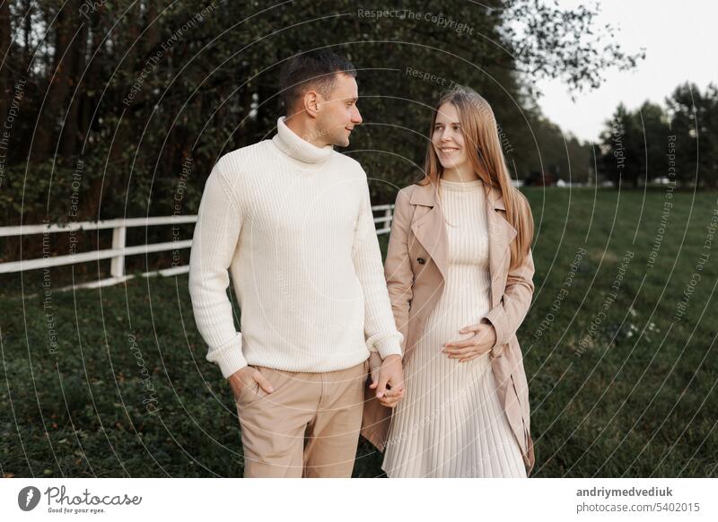 Young happy romantic pregnant couple is walking holding hands outdoors at autumn warm day. Pregnant woman in knit dress and her handsome man expecting a baby. Happy family day. Parenthood, motherhood