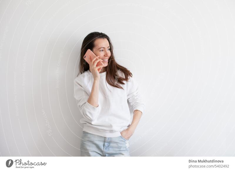 Young woman holding a mobile phone, smiling, using social media. Millennial person in white sweatshirt on white background. People using technology. Work call.