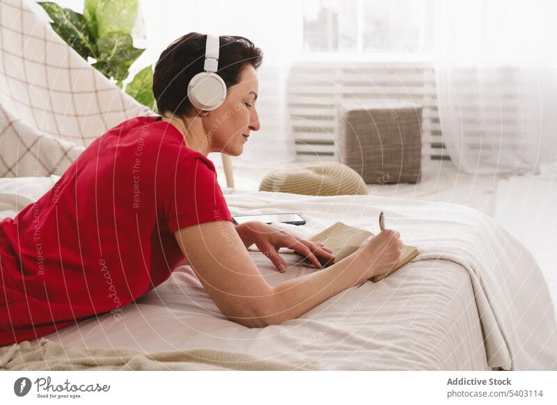 Woman listening to music and writing notes on bed woman focus concentrate write notebook take note headphones bedroom lying laptop female song home gadget