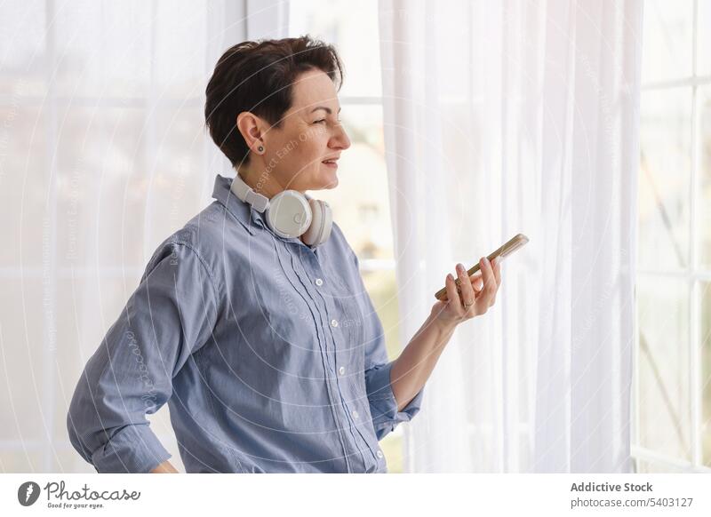 Mature woman holding smartphone while looking away headphones gadget device at home weekend using window studio style short hair female middle age mobile