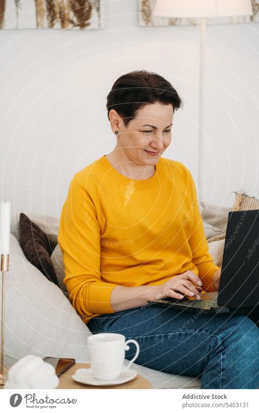 Smiling mature woman working on laptop from home smile positive freelance sofa gadget online at home coffee using remote cheerful device drink middle age female