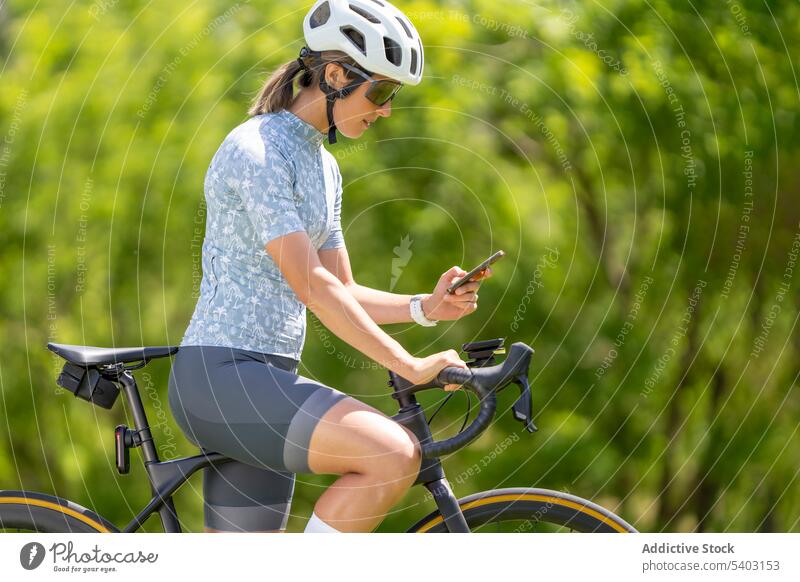 Woman on bicycle with smartphone against blurred park woman cyclist helmet sunglasses sportswear mobile browsing female bicyclist athlete racer safety hardhat