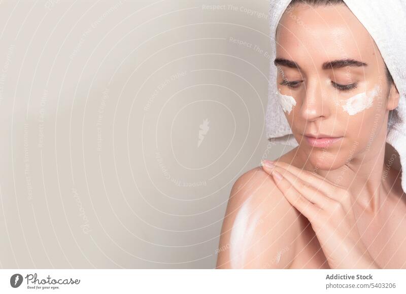 Serious young woman applying moisturizing cream on shoulder skin care smear moisture beauty routine towel cosmetic female procedure bare shoulders wall