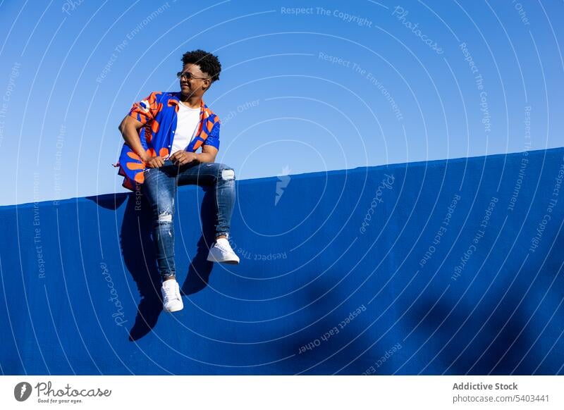 Stylish black man sitting on fence trendy confident concrete wall cool outfit relax fashion sneakers young male african american curly hair ethnic serious wear