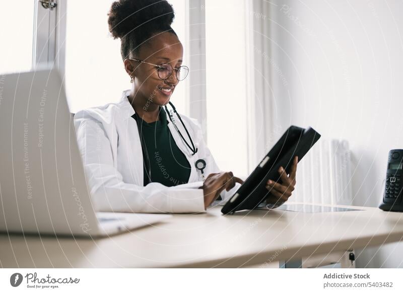 Smiling black woman sitting at table and browsing tablet doctor using smile uniform stethoscope specialist professional room female african american ethnic