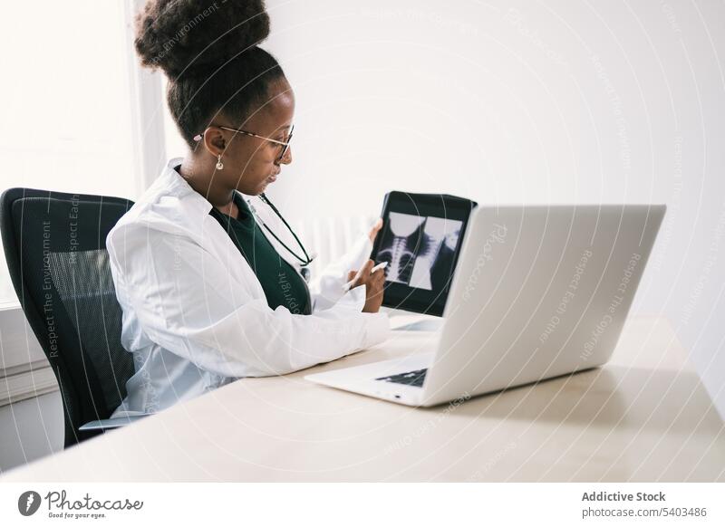 Side view of black doctor sitting at table and showing x ray images on tablet woman uniform stethoscope specialist professional room female ethnic