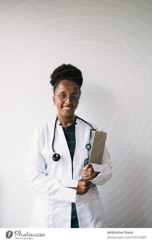 Happy ethnic doctor standing near wall with clipboard woman uniform positive smile professional stethoscope specialist young female happy friendly