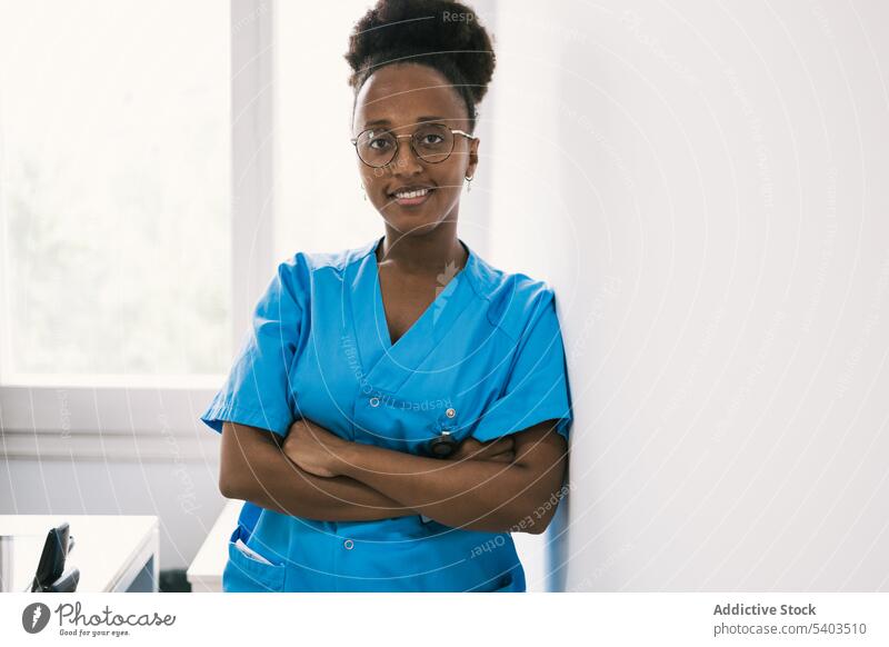 Smiling black woman in eyeglasses standing near wall doctor uniform positive smile hands crossed professional happy glad lean on young female african american