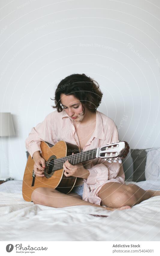 Serious young woman playing guitar on bed in bedroom music practice acoustic hobby leisure relax home female instrument guitarist melody comfort song talent