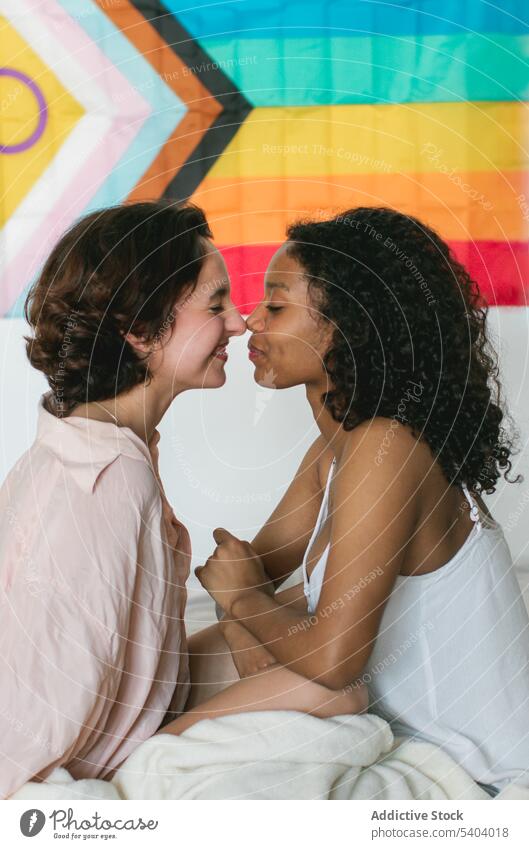 Happy diverse lesbian couple sitting together against rainbow flag women smile lgbt touch nose happy eyes closed blanket young female love homosexual