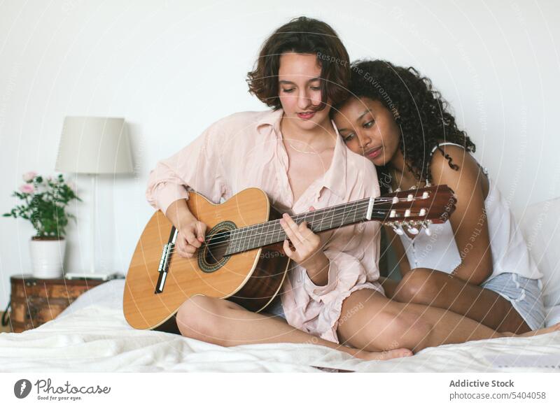Concentrated young caucasian woman playing guitar while african american girlfriend leaning head on shoulder and enjoying music lesbian couple leisure together