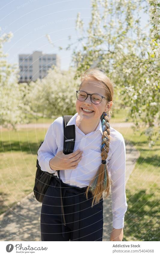 Smiling teenage girl standing with school bag in park schoolgirl cheerful carefree eyes closed smile positive adolescent sunlight happy eyeglasses style glad