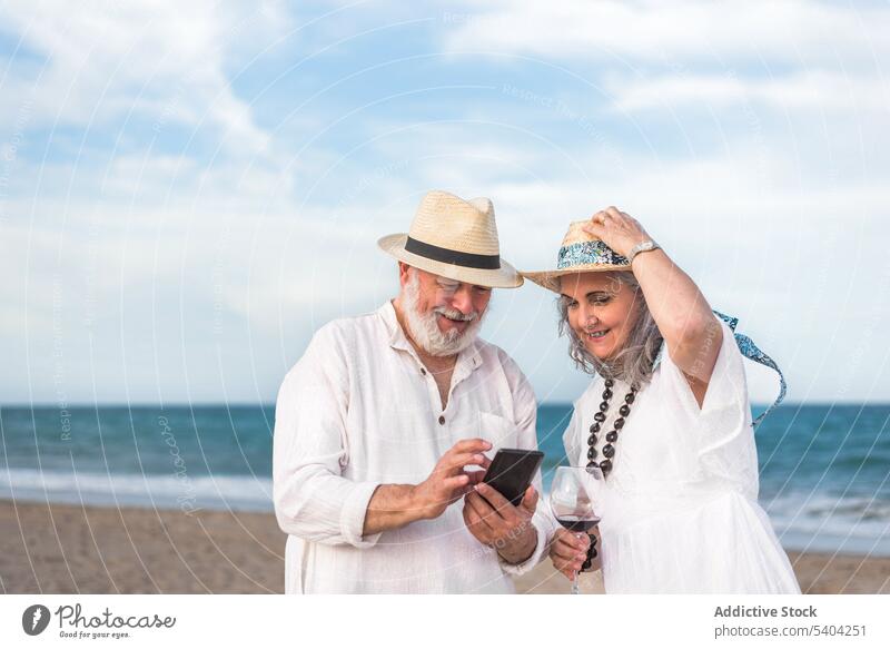 Senior couple using phone on sandy coast smartphone beach resort sea wind browsing summer device gadget straw hat surfing shore relationship tourism seashore