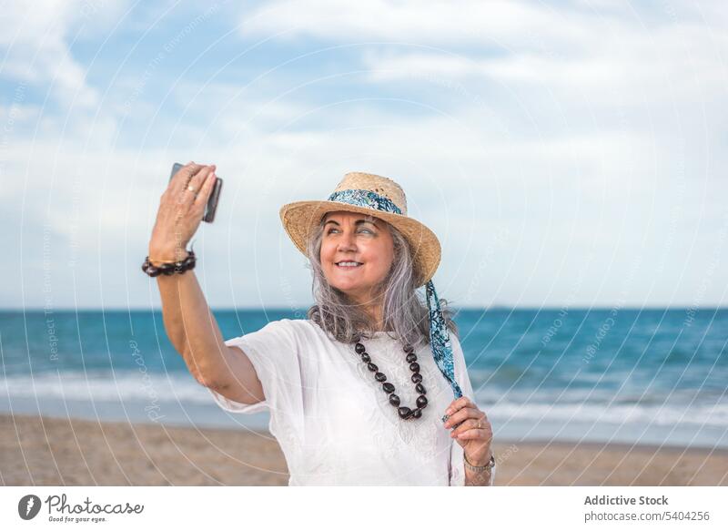 Mature woman in hat taking selfie on smartphone near sea using beach sand coast straw hat vacation summer mature female middle age gadget device mobile wave