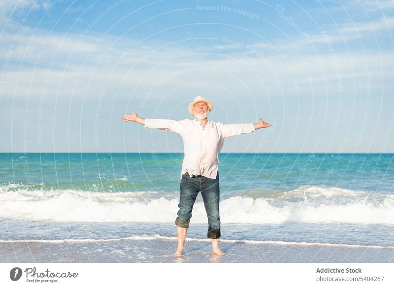 Middle aged man in hat standing on beach with outstretched arms straw hat eyes closed barefoot seawater recreation enjoy vacation male mature admire sand shore
