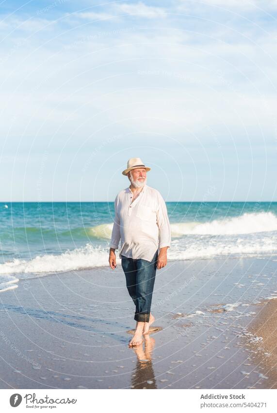 Mature man in hat walking along sandy beach near seawater shore barefoot straw hat cloudy blue sky vacation male mature middle age seashore summer coast wave