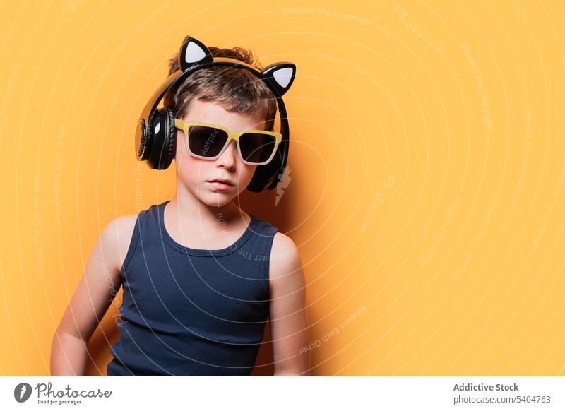 Stylish boy in sunglasses and headphones on yellow background trendy listen music using confident style wireless kid preteen child tank top shorts outfit bright