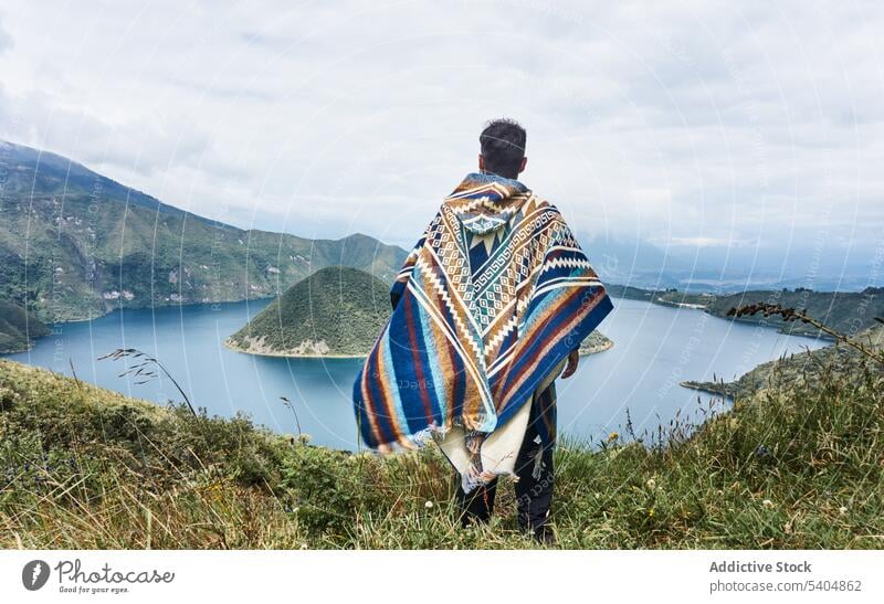 Unrecognizable man in warm clothes standing on grassy meadow and enjoying lake view traveler admire mountain highland nature landscape explore poncho male calm