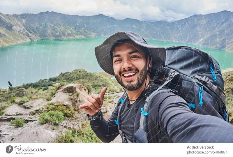 Cheerful man traveler with backpack and in hat standing on hill hiker selfie mountain smile trekking cheerful trip male young adventure highland nature gesture