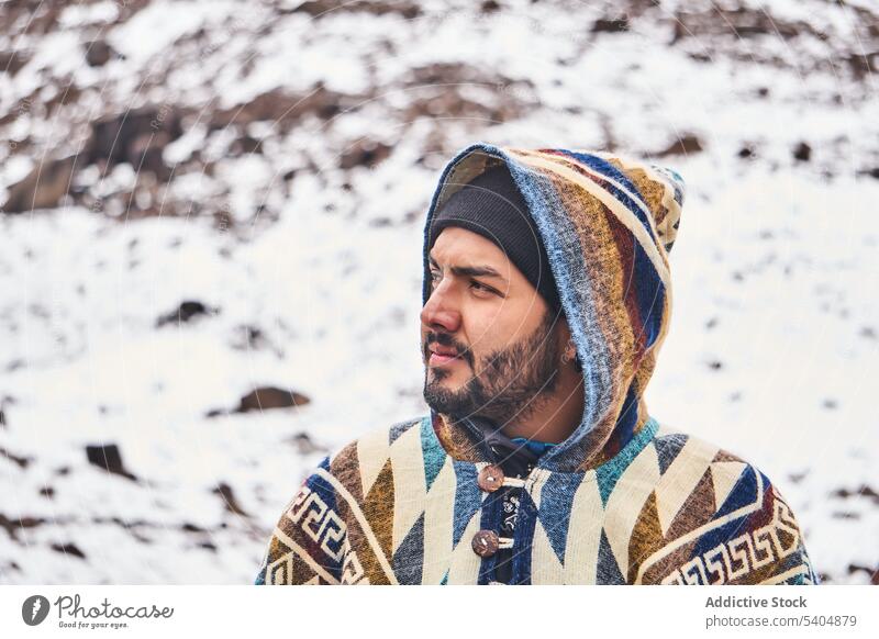 Pensive young man in cap and hood standing on snowy terrain portrait mountain cold winter poncho male beard nature outerwear warm wool style wintertime highland