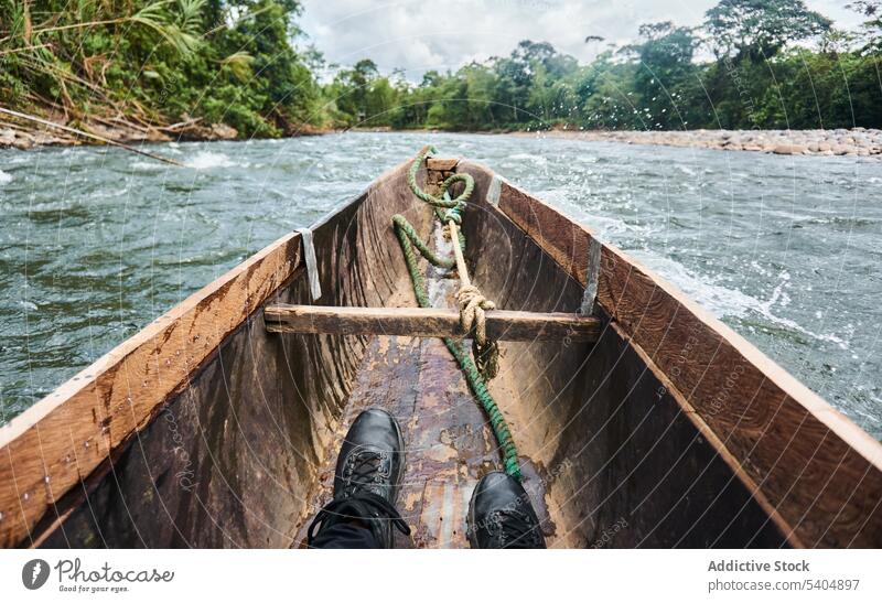 Anonymous man in black shoes sitting on traditional boat floating on river water tree wooden ride countryside rope male transport cloudy ripple tied coast bow