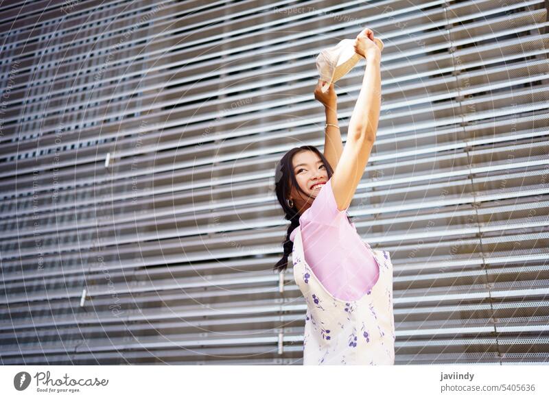 Carefree Asian woman against metal wall smile happy dance street arms raised carefree modern urban female young asian ethnic optimist energy glad cap style