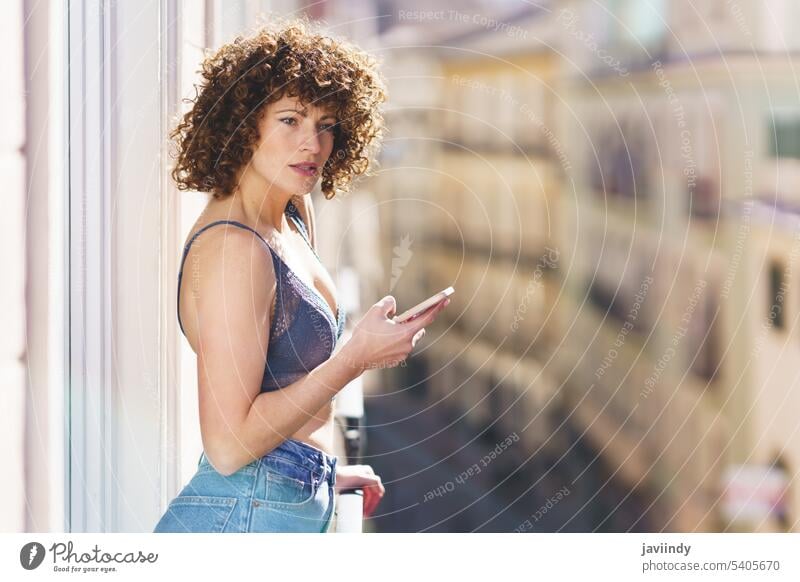 Young woman in bra and jeans with curly hair while browsing smartphone underwear hairstyle positive lingerie balcony summer rest sun home female young fashion