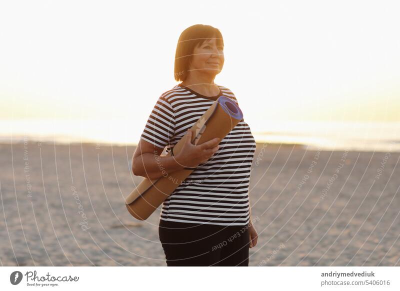 mature woman holding a sports mat and preparing to practice yoga outdoors on sea beach. Happy mature overweight woman exercising on seashore. copy space. Meditation, yoga and relaxation concept.