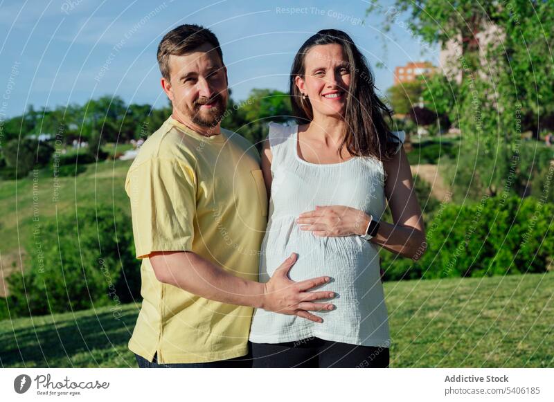 Happy couple in expectance embracing standing in park wife husband pregnant belly maternal embrace pregnancy await baby bump childbearing prenatal anticipate