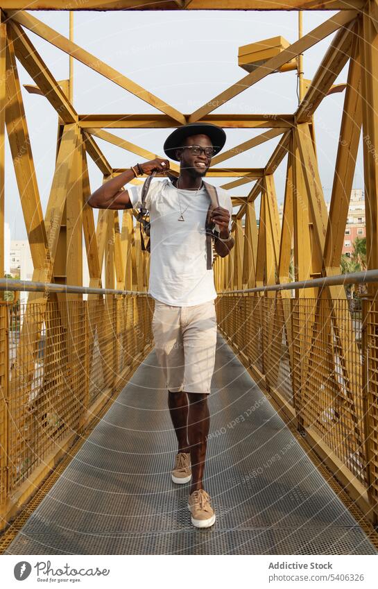 Cheerful black guy smiling while walking on footbridge in park man trendy smile tourist positive style city happy male young african american ethnic fashion hat