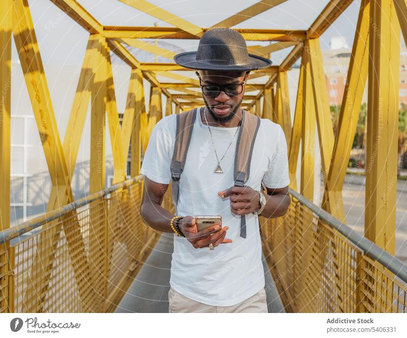 Trendy black male with phone walking on bridge man trendy tourist smartphone using stroll city street backpack device gadget mobile ethnic african american