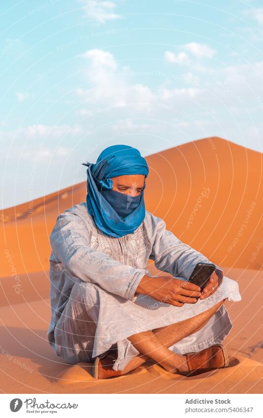Muslim man in traditional clothes using cellphone in desert berber smartphone sand dune tuareg browsing morocco merzouga blue turban sky cloud travel male