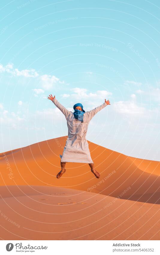Carefree Berber man jumping on sand in desert berber dune outstretch arms raised freedom tradition tuareg merzouga morocco sky summer sunny nature male journey