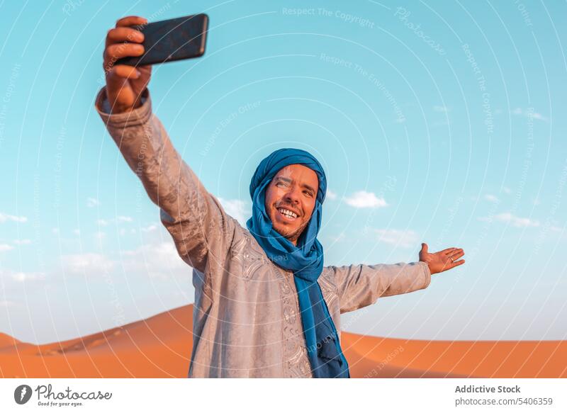 Cheerful Muslim man taking selfie in desert against blue sky berber using smartphone cheerful turban tradition smile tuareg muslim morocco travel take photo