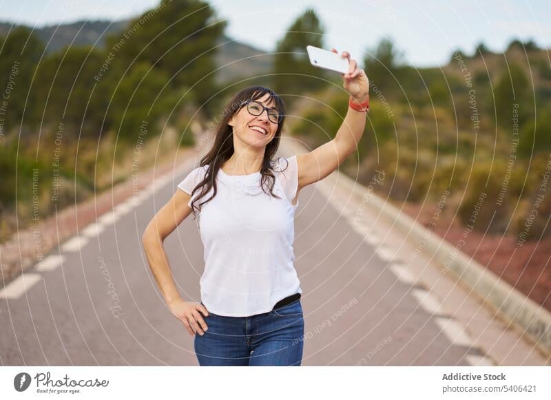 Cheerful woman with standing and taking selfie on road traveler smartphone tourist street take photo countryside vacation adult female gadget device mobile