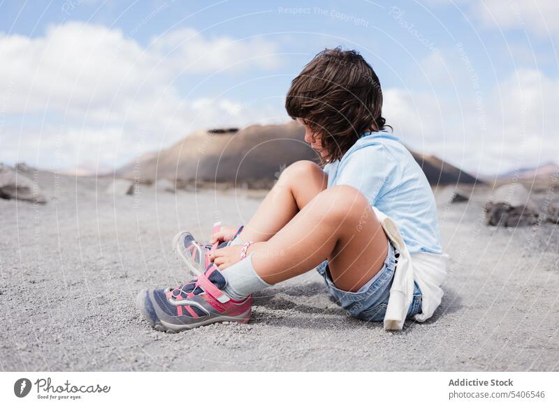 Unrecognizable child sitting on dry terrain and tying shoelaces kid tie sneakers ground activity summer blue sky countryside girl sunlight childhood preteen